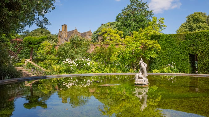 A large circular pond with a stone fountain in the centre and a garden with trees and a manicured hedge behind, an old manor house in the background.